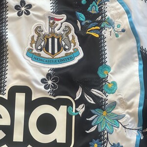 May include: White and black striped jersey with the Newcastle United crest and floral embroidery. The jersey features the team's logo, a crest with two seahorses, and the word "ela".