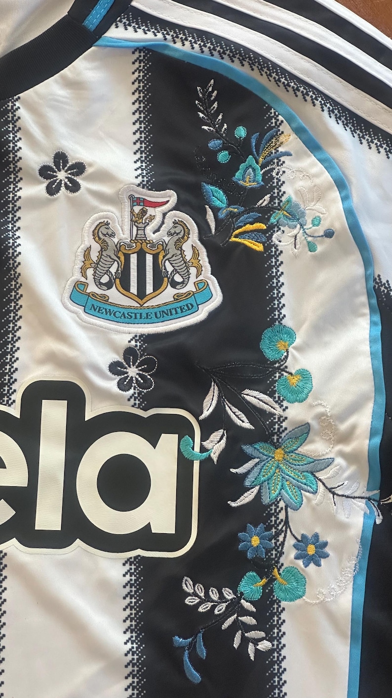 May include: White and black striped jersey with the Newcastle United crest and floral embroidery. The jersey features the team's logo, a crest with two seahorses, and the word "ela".