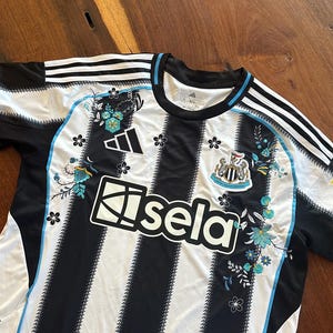 May include: Black and white striped soccer jersey with floral accents in turquoise and black. Features the Adidas logo, the Newcastle United crest, and the "sela" logo. The jersey has a blue trim.
