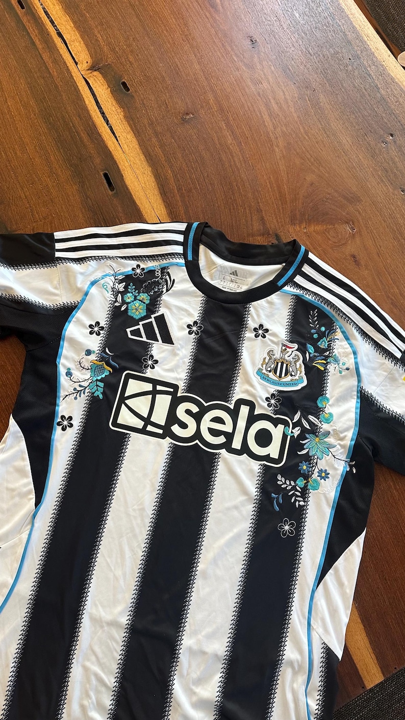 May include: Black and white striped soccer jersey with floral accents in turquoise and black. Features the Adidas logo, the Newcastle United crest, and the "sela" logo. The jersey has a blue trim.
