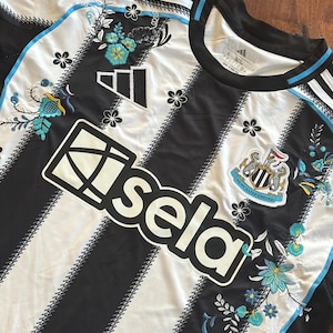 May include: Black and white striped soccer jersey with floral embroidery. Features the Adidas logo, the "Sela" logo, and the Newcastle United crest. The jersey has a crew neck and blue accents.