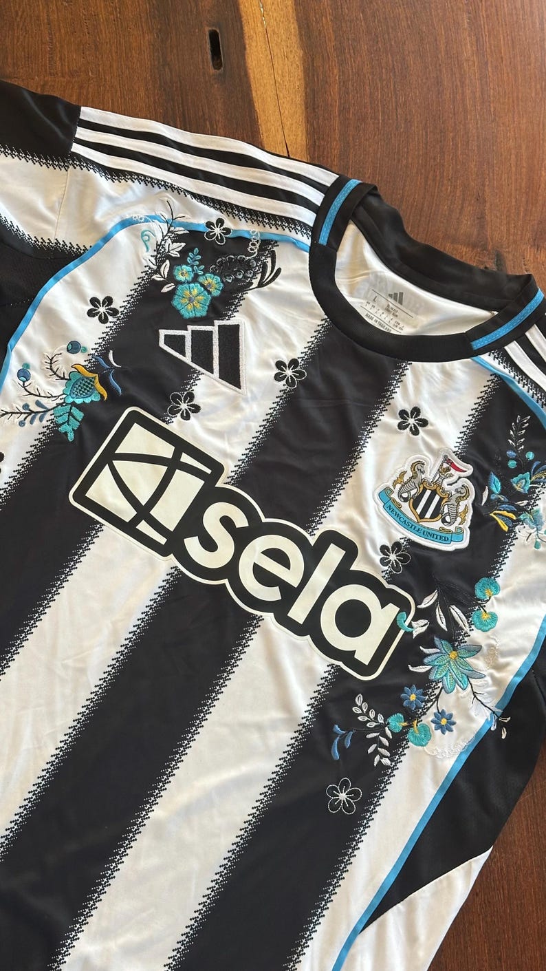 May include: Black and white striped soccer jersey with floral embroidery. Features the Adidas logo, the "Sela" logo, and the Newcastle United crest. The jersey has a crew neck and blue accents.