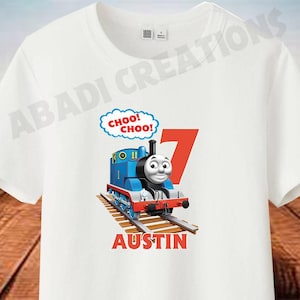 Puede incluir: Camiseta blanca con un diseño de Thomas la locomotora azul y rojo. El tren está en una vía con las palabras "Choo! Choo!" encima. El nombre "Austin" está impreso debajo del tren.