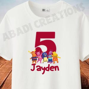 May include: White t-shirt with a large red number 5 and the name "Jayden" in red script. The shirt features a colourful graphic of cartoon characters. This is a children's apparel item.