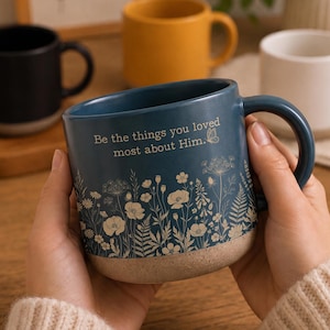 May include: A blue ceramic mug with a speckled beige base, featuring a white floral print and the text "Be the things you loved most about Him." with a small butterfly icon. The mug has a curved handle and a smooth, matte-finish surface.