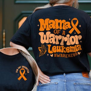 May include: Black t-shirt with orange text and a ribbon graphic. The text reads "Mama of a Warrior Leukemia Awareness" with a butterfly and heart. A smaller image on the front reads "For My Daughter" with a ribbon.