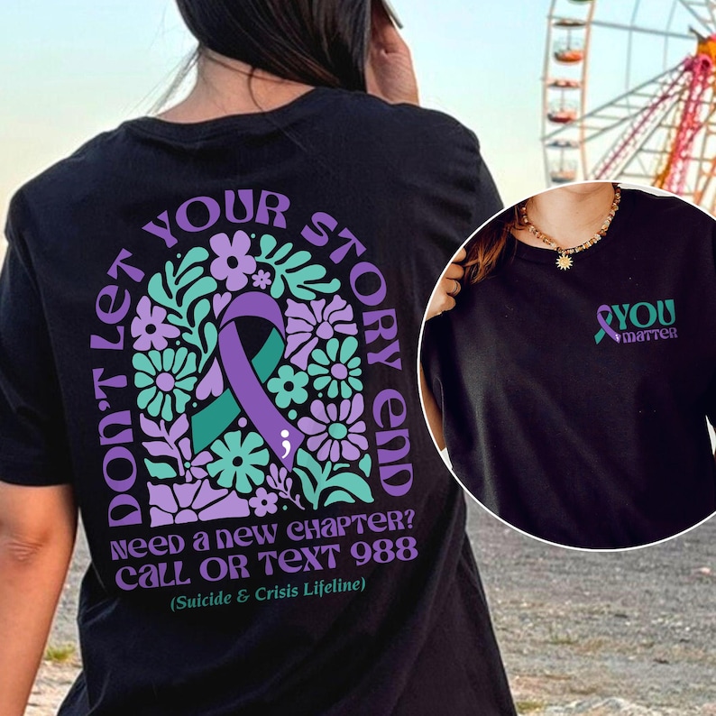 Puede incluir: Camiseta negra con una cinta morada y un dise&ntilde;o floral. El texto "Don't let your story end" est&aacute; escrito alrededor de la cinta. El texto "Need a new chapter? Call or text 988 (Suicide & Crisis Lifeline)" est&aacute; escrito debajo del dise&ntilde;o.