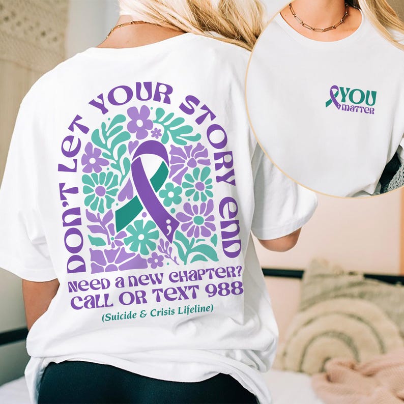 Puede incluir: Camiseta blanca con una cinta morada y un dise&ntilde;o floral en la espalda. El texto dice "No dejes que tu historia termine. &iquest;Necesitas un nuevo cap&iacute;tulo? Llama o env&iacute;a un mensaje de texto al 988 (L&iacute;nea de vida para el suicidio y la crisis)". La parte delantera de la camiseta tiene una cinta morada con el texto "T&uacute; importas".