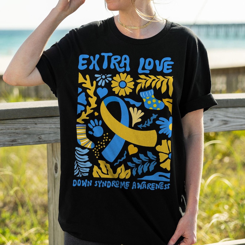Puede incluir: Camiseta negra con un dise&ntilde;o gr&aacute;fico colorido que presenta una cinta amarilla y azul, flores y el texto "Extra Love Down Syndrome Awareness".