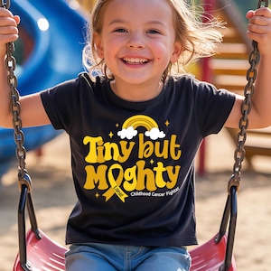 May include: A black t-shirt with the words "Tiny but Mighty" in yellow, featuring a rainbow and ribbon design. The shirt also includes the text "Childhood Cancer Fighter" and the name "Lanie". The shirt is worn by a child on a swing.