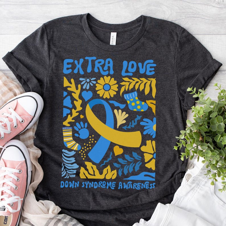 Puede incluir: Una camiseta gris oscuro con un dise&ntilde;o colorido que incluye una cinta amarilla y azul, flores y hojas. El texto "Extra Love Down Syndrome Awareness" est&aacute; impreso en la camiseta.