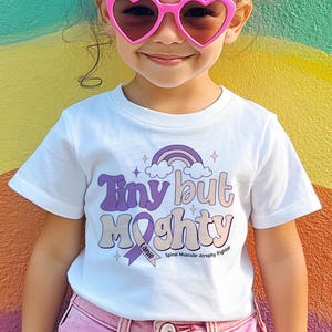 May include: A white t-shirt with the words "Tiny but Mighty" in purple and peach lettering, a rainbow graphic, and the text "Spinal Muscular Atrophy Fighter". The shirt is worn by a child wearing pink heart-shaped sunglasses.
