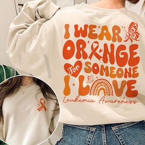 May include: A cream-colored sweatshirt with the text "I wear orange for someone I love Leukemia Awareness" in orange and red lettering. The sweatshirt features a heart with the word "for" inside, a butterfly, and a rainbow.