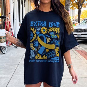 A Down Syndrome Awareness T-shirt with “3:21” and “Extra Love” printed in a heart-themed design. The unisex shirt represents support for the Down syndrome community and T21 families. Made of durable cotton with a relaxed fit, ideal for events, family gatherings, or spreading awareness every day.