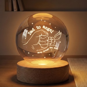 May include: A clear glass sphere with an illuminated design of two hands clasped together, with the words "Dad to angel" and "Baby Ortega Dec. 2025". The sphere sits on a round wooden base, emitting a warm glow.