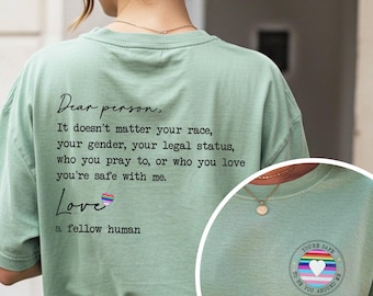 Puede incluir: Camiseta verde salvia con texto: "Querida persona, no importa tu raza, tu género, tu estatus legal, a quién rezas o a quién amas, estás a salvo conmigo. Amor, un ser humano". También hay un pequeño gráfico.