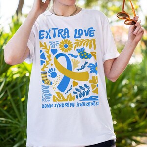 A cotton T-shirt featuring “T21 Mama” with a Down Syndrome Awareness ribbon and “Extra Love” graphic. The unisex shirt celebrates families and supporters of Down syndrome awareness. It’s soft, comfortable, and perfect for awareness events, daily wear, or family photos celebrating inclusion.