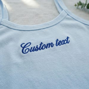 May include: Light blue tank top with thin straps. The words "Custom text" are embroidered in a dark blue script across the chest. The top is made of a soft material.