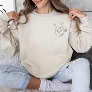 Pu&ograve; includere: Una felpa beige con una grafica di una mano che fa il segno 'Ti amo' nella Lingua dei Segni Italiana. Il testo 'L'amore non ha bisogno di parole' &egrave; sotto la grafica della mano.