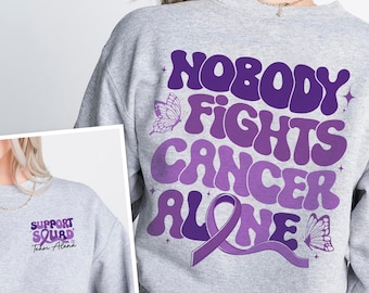 May include: Grey sweatshirt with a purple and white design that says "Nobody Fights Cancer Alone" with a purple ribbon and butterflies. The front of the sweatshirt has a smaller design that says "Support Squad Team Alana".