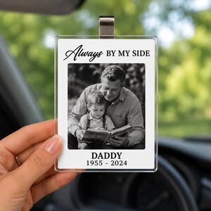 May include: A clear, rectangular car charm with a metal clip. The charm features a black and white photo of a man and child reading a book. Text on the charm reads "Always BY MY SIDE" and "DADDY 1955 - 2024".