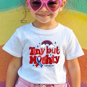 May include: White t-shirt with the text "Tiny but Mighty" in red and blue, featuring a rainbow and stars graphic. The shirt also includes the text "Lanie" and "CHD Warrior".