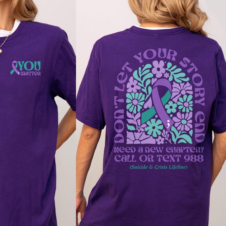 Puede incluir: Camiseta morada con un lazo morado y un dise&ntilde;o floral en la espalda. El texto en la espalda dice "No dejes que tu historia termine. &iquest;Necesitas un nuevo cap&iacute;tulo? Llama o env&iacute;a un mensaje de texto al 988 (L&iacute;nea de vida para el suicidio y la crisis)". La parte delantera de la camiseta tiene un lazo morado y el texto "T&uacute; importas".