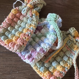 Puede incluir: Bolsas de crochet hechas a mano en colores pastel. Las bolsas presentan un patrón de puntada texturizada, similar a burbujas. La paleta de colores incluye rosas, amarillos, verdes y azules suaves. Las bolsas se utilizan probablemente para objetos pequeños o regalos.