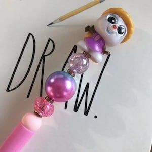 May include: A pink pen with a decorative beaded design and a small cartoon character on top. The beads are various colors, including pink, purple, and clear. The word "DRAW" is written in black on the white background.