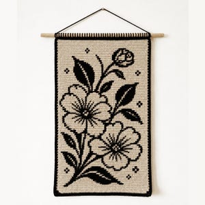 May include: A rectangular crochet wall hanging featuring a beige background with a black floral pattern. The design displays two large blossoms, a small bud, and several leaves. It is suspended from a light wooden dowel with a black cord.