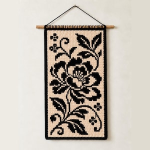 Puede incluir: Un tapiz rectangular con un patrón floral de punto de cruz negro sobre un fondo de tela beige. La pieza está suspendida de un pasador de madera con una cuerda negra, mostrando una gran flor central con hojas y pequeños capullos.