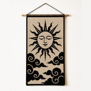 May include: A rectangular crochet wall hanging featuring a black and beige sun with a serene face and stylized rays above black swirling clouds. The textile is suspended from a horizontal wooden dowel with a black cord for wall mounting.