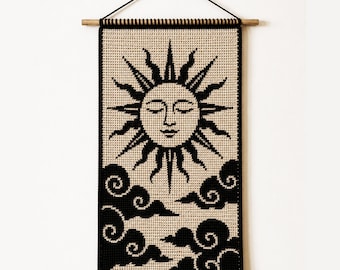 May include: A rectangular crochet wall hanging featuring a black and beige sun with a serene face and stylized rays above black swirling clouds. The textile is suspended from a horizontal wooden dowel with a black cord for wall mounting.