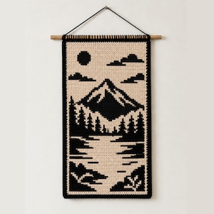 Op de afbeelding: Een rechthoekige wandhanger met een zwart en beige kruissteekpatroon van een berglandschap. Het ontwerp toont een centrale bergtop, een dennenbos, een meer, wolken en een zon, opgehangen aan een houten roede.