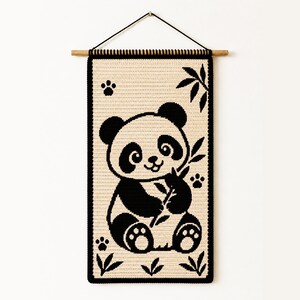 Pode incluir: Uma tapeçaria retangular de crochê com um design de urso panda em preto e creme. O panda é retratado segurando um caule de bambu, cercado por pequenas pegadas e folhas de bambu. Está suspenso por um suporte de madeira com um cordão preto.