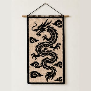 Peut inclure: Une tenture murale rectangulaire présentant un motif de dragon tricoté en noir sur un fond beige. La pièce est suspendue à une tige en bois avec un cordon noir. Le motif du dragon est entouré de petites formes de nuages noirs stylisés.