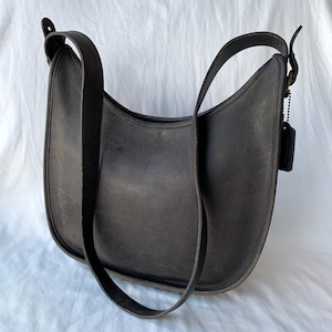 May include: A dark gray leather shoulder bag with a curved design. The bag has a long, adjustable strap and a small tag attached to a chain. The leather appears worn, giving it a vintage look. The bag is set against a white background.