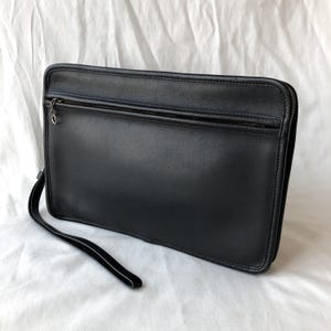 Könnte beinhalten: Eine schwarze Leder-Clutch mit Reißverschluss und Handschlaufe. Die rechteckige Tasche hat eine glatte Oberfläche und einen silberfarbenen Reißverschluss. Die Handschlaufe ist an der Seite der Tasche befestigt.