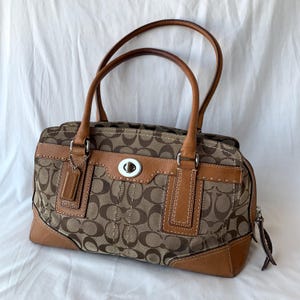 May include: A tan and brown handbag featuring a repeating C logo pattern on canvas fabric. The bag includes tan leather trim, two top handles, a silver-tone metal turn-lock closure on the front, and a small leather luggage tag attached to a handle.