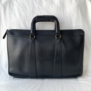 Op de afbeelding: Een zwarte leren aktetas met een rechthoekig silhouet, een ritssluiting aan de bovenkant en twee gestructureerde handvatten bevestigd met messingkleurige hardware. De tas heeft een gladde afwerking met verticale stiksels vanaf de handvatten.