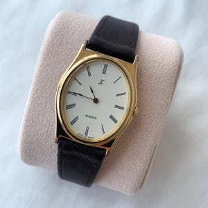 Può includere: Un orologio da polso color oro con quadrante ovale bianco, numeri romani neri e lancette sottili nere. L'orologio presenta un cinturino in pelle marrone scuro e le scritte JF, QUARTZ e JAPAN 1300-6A00 R stampate sul quadrante.