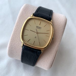 May include: A gold-tone Seiko Quartz wristwatch with a square-shaped case and a textured gold dial. The watch features thin gold-tone hour markers, three central hands, and a black leather strap with a visible grain texture and a metal buckle.
