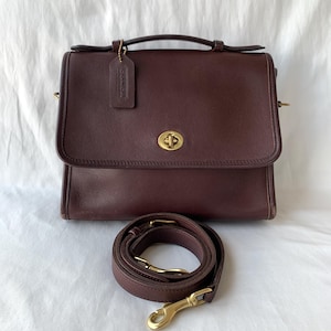 Op de afbeelding: Een donkerbruine leren tas met een handvat aan de bovenkant, een voorflap met een messing draaisluiting en een bijpassende afneembare schouderriem met messing draaihaken. Een leren Coach-merklabel hangt aan het handvat met een klein kettinkje.