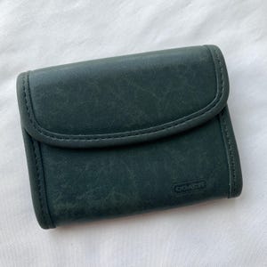 May include: A dark green leather wallet with a flap closure. The wallet has a stitched border and a small embossed "COACH" logo. The leather has a slightly distressed appearance.