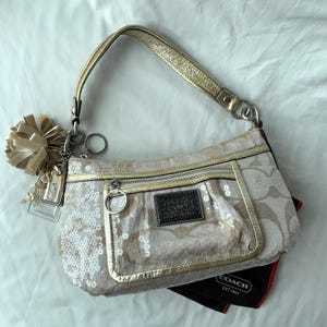 May include: A beige and gold Coach shoulder bag featuring a signature monogram pattern, white sequin embellishments, a metallic gold leather strap, a front zippered pocket, a dark brown leather logo patch, and a decorative gold pom-pom charm.