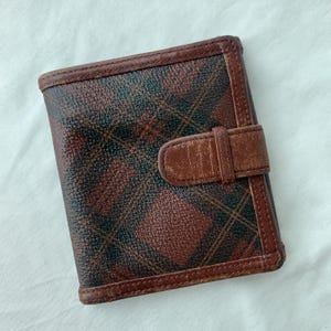 May include: A brown and black plaid wallet with a leather strap closure. The wallet has a rectangular shape with brown leather trim and a textured surface. The plaid pattern features red, black, and brown colors.