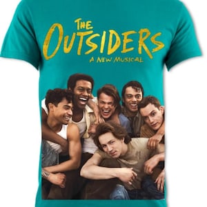 May include: Teal t-shirt featuring the musical title "The Outsiders" in yellow distressed lettering. Below the title is the text "A New Musical". The shirt displays a group of smiling men. A casual tee for fans of the musical.