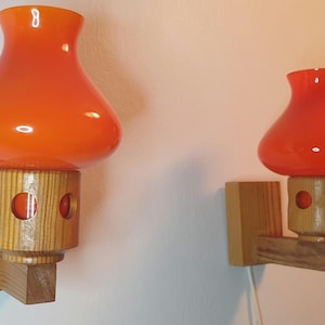 Può includere: Due applique da parete in vetro arancione con basi in legno. Le applique hanno una forma rotonda e bombata e sono montate su una parete.