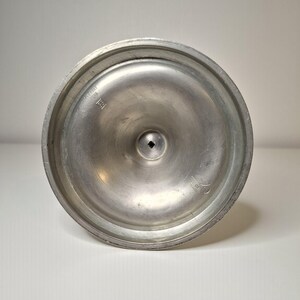 K&ouml;nnte beinhalten: Nahaufnahme der Unterseite eines silberfarbenen Metallobjekts. Das Objekt hat eine kreisf&ouml;rmige Basis mit einem erh&ouml;hten Rand und einem kleinen quadratischen Loch in der Mitte. Auf der Basis befinden sich Markierungen, darunter ein Herstellerzeichen.