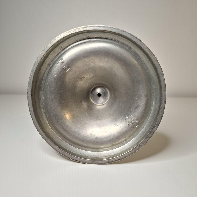 K&ouml;nnte beinhalten: Nahaufnahme der Unterseite eines silberfarbenen Metallobjekts. Das Objekt hat eine kreisf&ouml;rmige Basis mit einem erh&ouml;hten Rand und einem kleinen quadratischen Loch in der Mitte. Auf der Basis befinden sich Markierungen, darunter ein Herstellerzeichen.